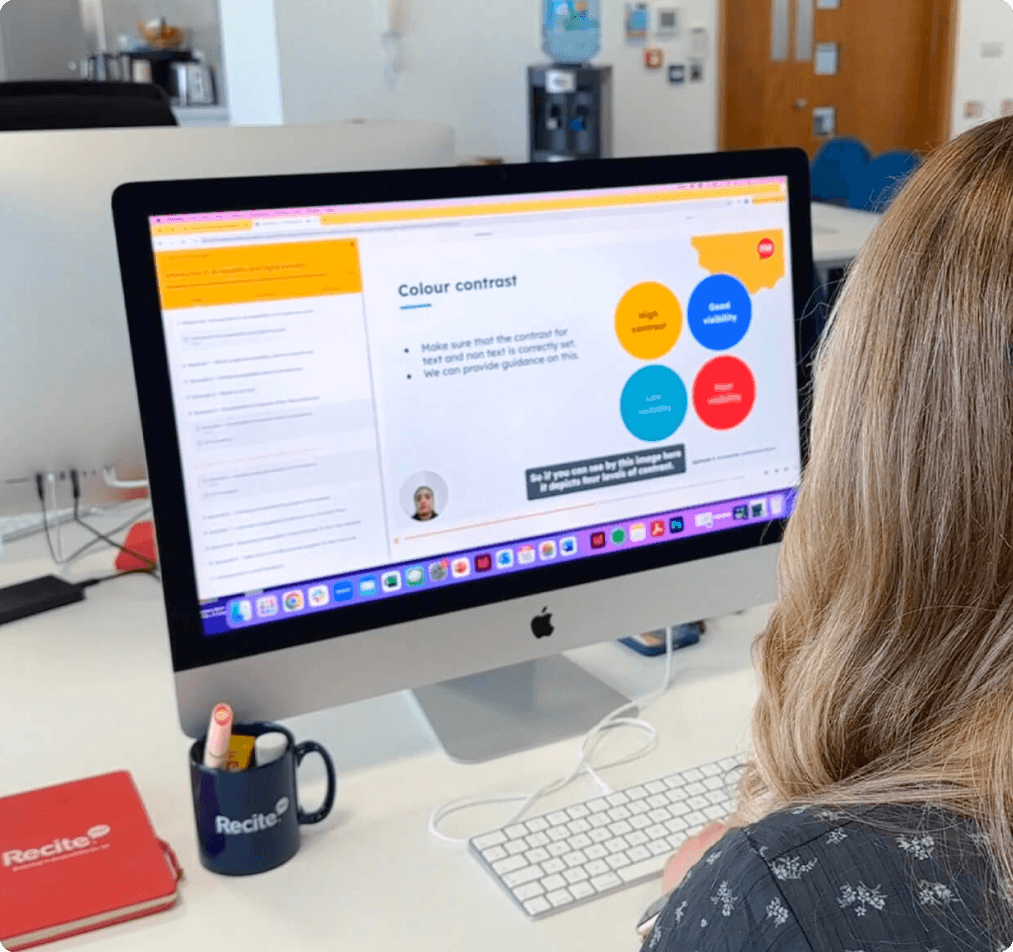Person with long hair sitting at a desk, viewing a presentation on an iMac screen about colour contrast in accessibility. The slide shows four colored circles labeled high contrast, good visibility, low visibility, and poor visibility. On the desk are a Recite-branded mug with pens and a red Recite notebook.