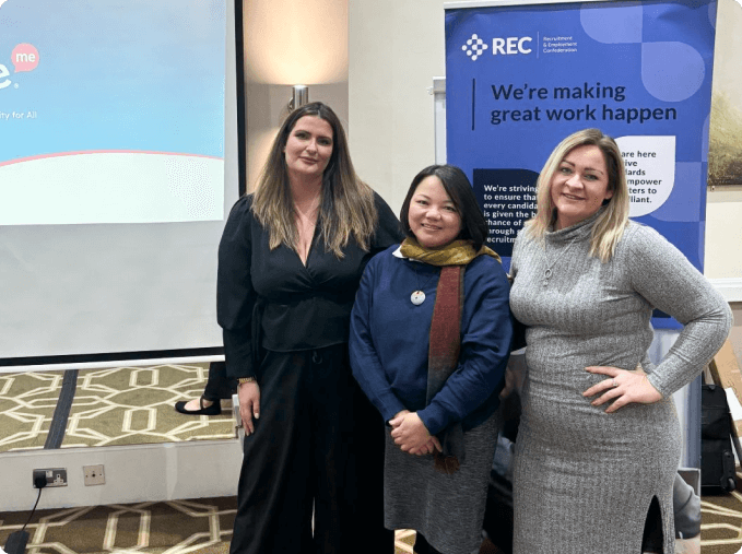 Three women stand together and smile at a professional event. Behind them is a display banner for the Recruitment and Employment Confederation (REC) with the slogan "We're making great work happen," and a projection screen showing part of a presentation.